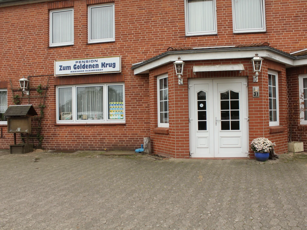 Backsteingebäude mit weißen Türen und Fenstern, über denen ein Schild "Pension Zum Goldenen Krug" prangt.