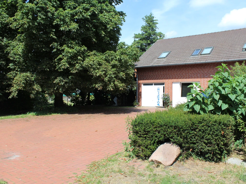 Kontraste, Diepenau  Backsteingebäude mit Satteldach und Dachfenstern, umgeben von dichter Vegetation und Pflastersteinen.
