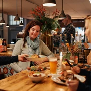 Johann Schäfer Brewery Zwei Frauen sitzen an einem naturbelassenem Holztisch, in einem gemütlich warm beleuchteten Restaurant. Eine Kellnerin serviert einen Teller.Two women are sitting at a natural wooden table in a cozy, warmly lit restaurant. A waitress is serving a plate.