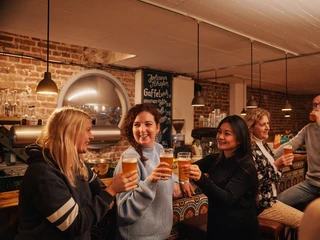 Johann Schäfer Brewery Drei Frauen stehen an der Bar des Restaurants und stoßen mit einem Bier an. Three women are standing at the bar of a restaurant and toasting with a beer.