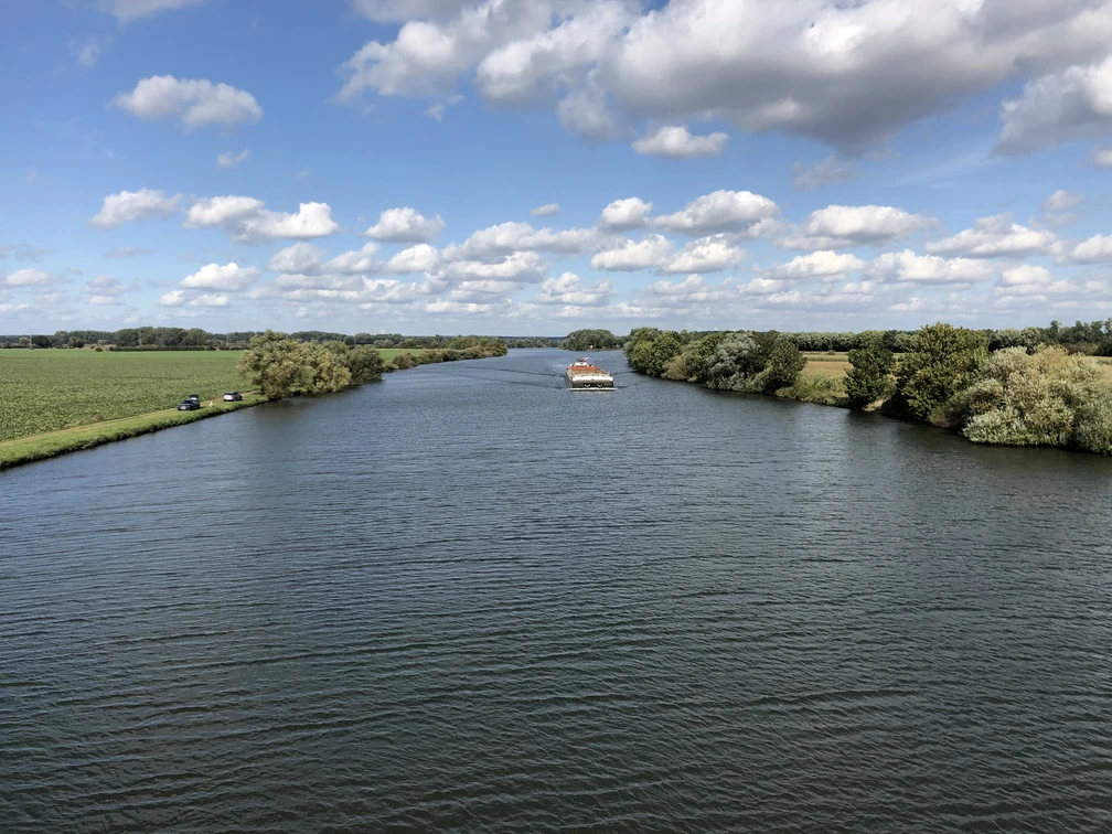 Breitflächiger Fluss, gesäumt von üppigem Grün, unter strahlend blauem Himmel mit ziehenden Wolken.