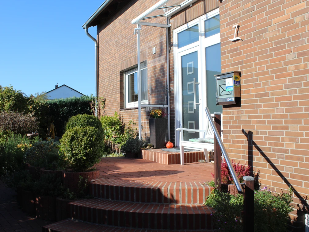 Eingang eines Backsteinhauses mit gläsernem Vordach und Treppe, umgeben von Pflanzen und Blumenschmuck.