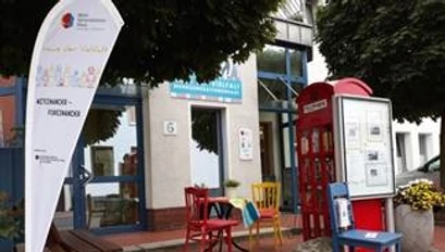MGH.jpg Ein Treffpunkt mit bunten Stühlen und Tischen vor dem Mehrgenerationenhaus und einer roten Telefonzelle.