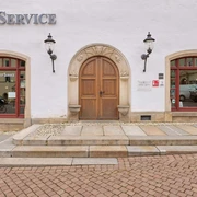 Canalettohaus Pirna Weiße Fassade mit brauner Holztür, flankiert von zwei roten Fenstern mit "Tourist Service"-Schriftzug, und zwei Laternen an der Wand.