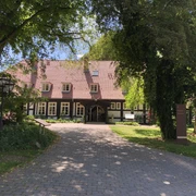 Ein Landhaus mit roten Dachziegeln und großen Fenstern, umgeben von grünen Bäumen und gepflegtem Rasen.