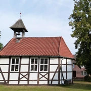 Historische Fachwerkkirche mit Glockenturm und rotem Ziegeldach, umgeben von grünem Rasen und Bäumen.
