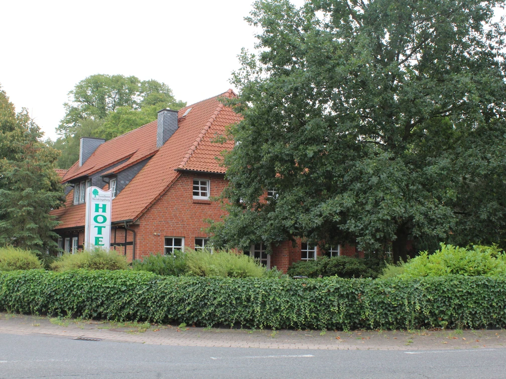 Landhaus Meinkingsburg Linsburg  Hotelgebäude mit rotem Ziegeldach, grüner Hecke und Bäumen im Vordergrund an einer Straßenecke.