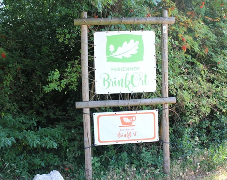 Ferienhof BrinkOrt, Warmsen  Eingangsschild des Ferienhofs BrinkOrt Warmsen, umgeben von üppigem, grünem Blätterwerk.
