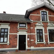 Historische rote Backsteinvilla mit weißen Fensterrahmen und hölzernen Türen, umgeben von Bäumen.