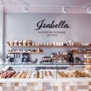 Isabella Glutenfreie Patisserie Blick auf eine Theke, hinter deren Glas kleine Gepäckstücke mit Beeren sowie Brotwaren ausgelegt sind. Auf der Wand dahinter steht "Isabella Glutenfreie Patisserie seit 2014" in Großbuchstaben. Darunter ist ein langes Holzbrett an der Wand befestigt, auf dem Gläser, Tassen, Gewürze sowie eine Topfpflanze stehen. Auf zwei kleineren Holzbrettern darunter stehen weitere Tassen und gestapelte Untertassen. Auf dem Küchentisch darunter steht eine große Kaffeemaschine sowie weitere Küchenutensilien.