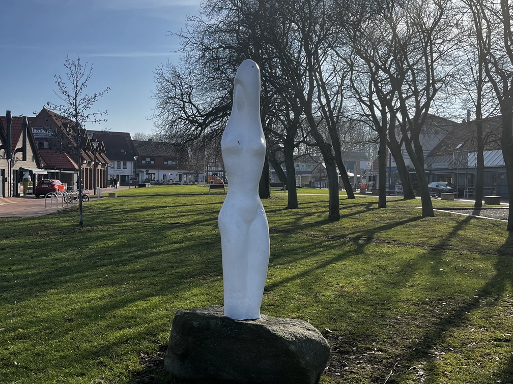 Eine weiße, abstrakte Statue steht auf einem grünen Rasen, umgeben von kahlen Bäumen und Häusern.
