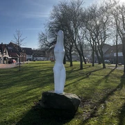 Eine weiße, abstrakte Statue steht auf einem grünen Rasen, umgeben von kahlen Bäumen und Häusern.