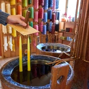 Kerzen selber ziehen  Eine Hand taucht zwei gelbe Kerzen in flüssiges Wachs; im Hintergrund sind bunte Kerzen an der Wand ausgestellt.