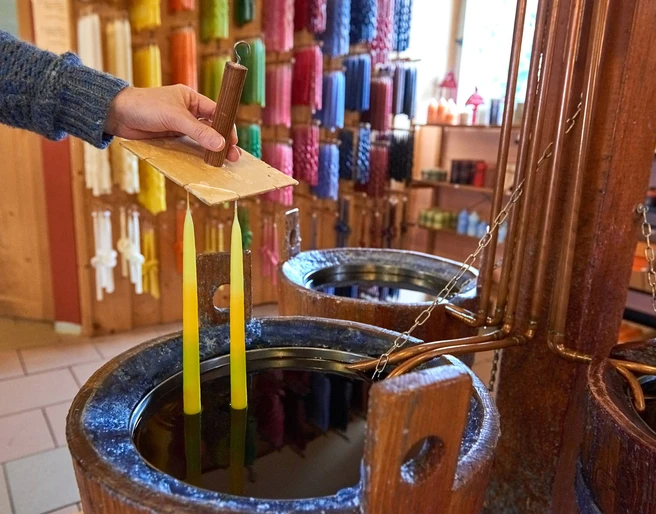 Kerzen selber ziehen  Eine Hand taucht zwei gelbe Kerzen in flüssiges Wachs; im Hintergrund sind bunte Kerzen an der Wand ausgestellt.