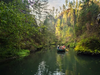 Obere Schleuse Hinterhermsdorf Eine Gruppe von Menschen fährt in einem Boot auf einem ruhigen Fluss, umgeben von dichtem, grünem Wald und steilen Felswänden.