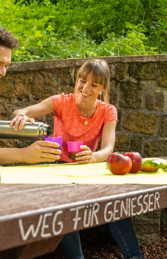 Werther-Arminiusquelle-Teutoburger-Wald-Tourismus-D-Ketz-077.jpg Ein Paar genießt ein Picknick an einem Holztisch im Grünen. Auf dem Tisch liegen Äpfel und Getränke.