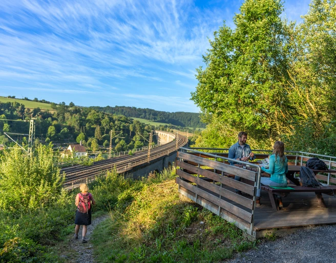 Altenbeken-Viadukt-Teutoburger-Wald-Tourismus-Patrick-Gawandtka-007.jpg Zwei Personen sitzen an einem Tisch mit Blick auf den Altenbekener Viadukt im Grünen. Eine Wanderin läuft einen Pfad entlang.
