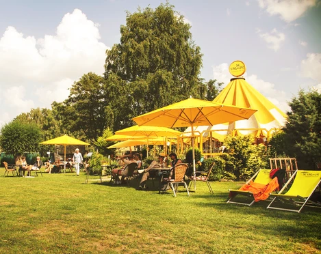 Schirmbar (Jens Butz).jpg Blick auf eine Wiese mit mehreren Sitzplätzen, gelben Sonnenschirmen und Menschen, die dort zusammensitzen und sich unterhalten