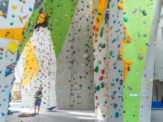 YOYO Kletterhalle  Kletterhalle mit bunten Kletterwänden; eine Person klettert, während eine andere sichert. Helle, lebendige Atmosphäre.