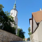 Stadtkirche Dohna Kirchturm mit grünem Dach und Wetterfahne ragt über Bäume und Kopfsteinpflasterstraße, flankiert von alten Gebäuden, unter klarem, blauem Himmel.