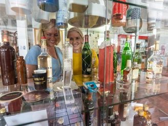 Steinhagen-HistorischesMuseum-Teutoburger-Wald-Tourismus-Patrick-Gawandtka-036.jpg Zwei Frauen betrachten Flaschen in einem Regal, umgeben von verschiedenen Spirituosen in einer Ausstellung.