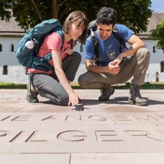 Höxter-Brenkhausen-Kloster-Teutoburger-Wald-Tourismus-A-Röser-042.jpg Zwei Wanderer betrachten Schriftzüge auf einer Steinplatte vor einem historischen Klostergebäude.