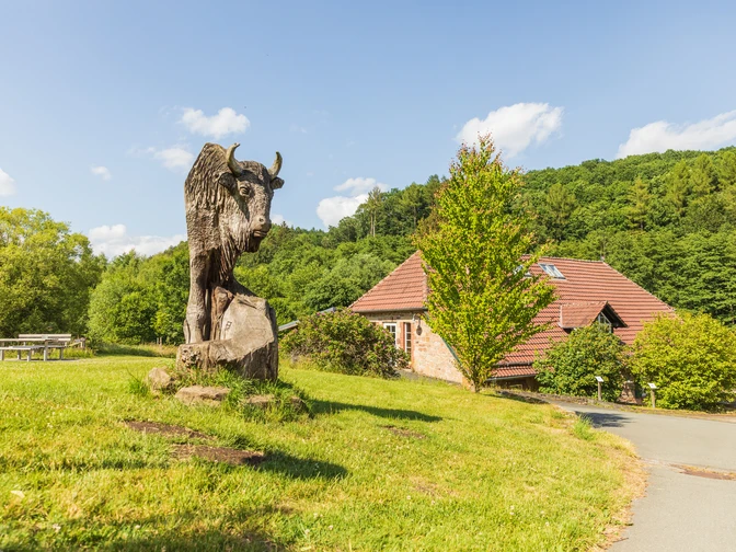 Steinbüffel-Skulptur auf grüner Wiese, Holzhütte und bewaldeter Hügel im Hintergrund.