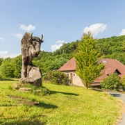 Warburg-Hammerhof-Teutoburger-Wald-Tourismus-A-Röser-051-CC-BY-SA.jpg Steinbüffel-Skulptur auf grüner Wiese, Holzhütte und bewaldeter Hügel im Hintergrund.