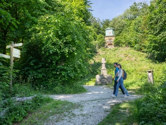 Halle-HagedornDenkmal-Teutoburger-Wald-Tourismus-Patrick-Gawandtka-087.jpg Ein Paar spaziert auf einem Waldweg vorbei an einem neoklassischen Denkmal im sommerlichen Grün.