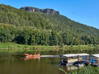 Kanu Aktiv Tours Ein orangefarbenes Motorboot fährt auf einem Fluss, flankiert von grünen Bäumen und einem bewaldeten Hügel im Hintergrund unter klarem, blauem Himmel.