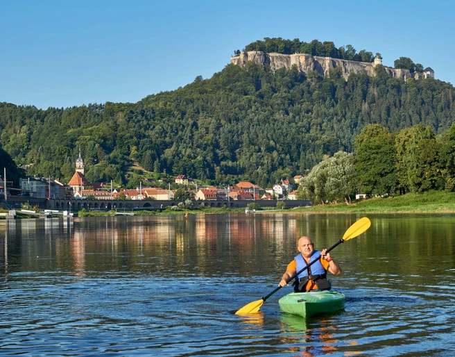 Kanu Aktiv Tours Ein Mann paddelt in einem grünen Kajak auf einem Fluss, im Hintergrund eine Stadt mit Kirche und bewaldetem Hügel unter klarem Himmel.