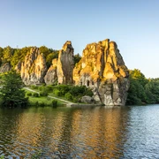 Felsenformation in grüner Landschaft spiegelt sich im See, umgeben von Bäumen bei Sonnenuntergang.