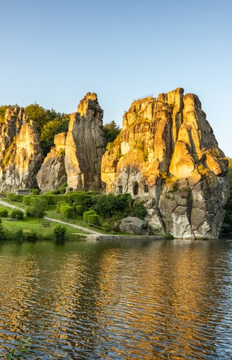 Horn-Bad Meinberg-Externsteine-Teutoburger-Wald-Tourismus-D-Ketz-010-CC-BY-SA.jpg Felsenformation in grüner Landschaft spiegelt sich im See, umgeben von Bäumen bei Sonnenuntergang.