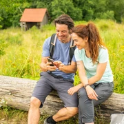 Horn-Bad Meinberg-Preußischer Velmerstot-Teutoburger-Wald-Tourismus-D-Ketz-088.jpg Ein lächelndes Paar in Wanderausrüstung sitzt auf einem Baumstamm und schaut gemeinsam auf ein Handy.