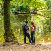 Detmold-Donoper Teich-Pivitker Wasserweg-Teutoburger-Wald-Tourismus-D-Ketz-089.jpg Zwei Wanderer stehen am Ufer eines idyllischen Waldbachs, umgeben von üppigem Grün der Natur.