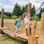 Bildungsstätte Sellnitz Zwei Kinder balancieren auf einem hölzernen Kletterparcours im Freien, umgeben von Bäumen und einer Wiese unter blauem Himmel.