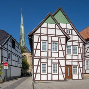 Fachwerkhäuser vor blauem Himmel mit Kirchturm im Hintergrund, gepflasterte Straße ohne Verkehr.
