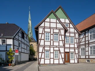 Fachwerkhäuser vor blauem Himmel mit Kirchturm im Hintergrund, gepflasterte Straße ohne Verkehr.