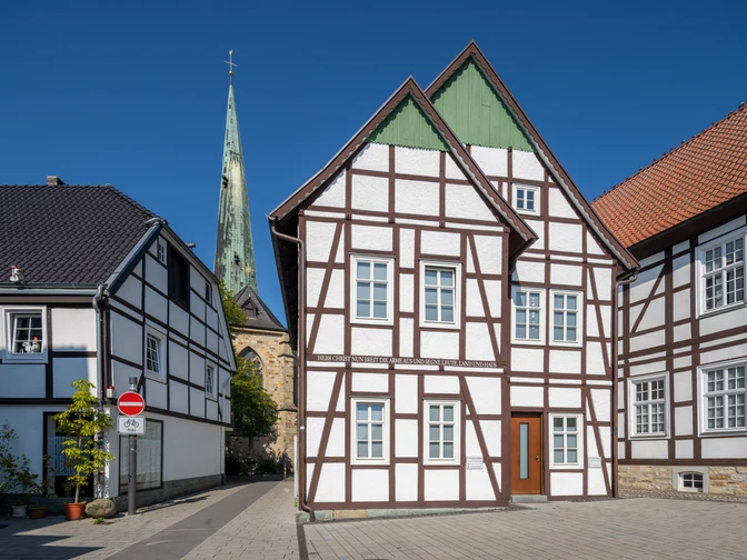 Fachwerkhäuser vor blauem Himmel mit Kirchturm im Hintergrund, gepflasterte Straße ohne Verkehr.