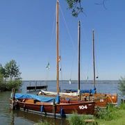Auswanderer am Ufer Drei traditionelle hölzerne Segelboote liegen vertäut an einem ruhigen Seeufer, umgeben von grünem Schilf.