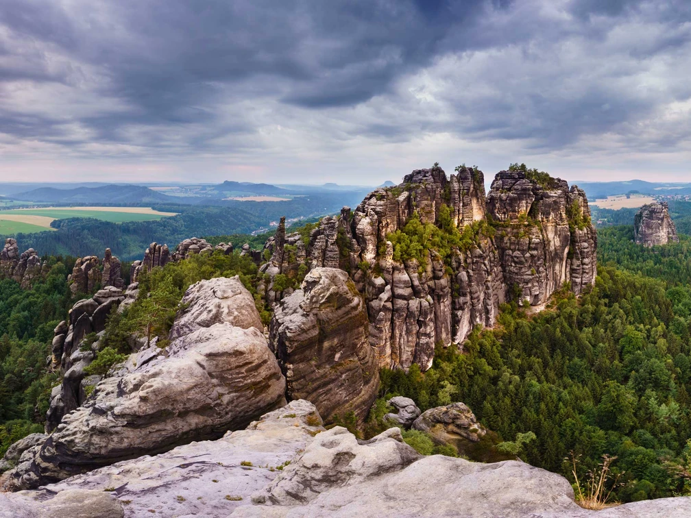 View from the Schrammstein lookout Felsformationen der Schrammsteine in der Sächsischen Schweiz, umgeben von dichtem Wald unter einem bewölkten Himmel, mit Blick auf eine weite Landschaft.