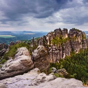 View from the Schrammstein lookout Felsformationen der Schrammsteine in der Sächsischen Schweiz, umgeben von dichtem Wald unter einem bewölkten Himmel, mit Blick auf eine weite Landschaft.