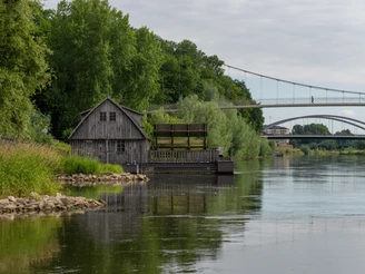 Minden-Schiffmühle-Teutoburger-Wald-Tourismus-Patrick-Gawandtka-029-CC-BY-SA.jpg Historische Wassermühle an der Weser bei Minden, umgeben von grünen Bäumen unter grauem Himmel.
