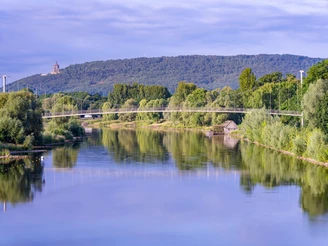 Ein idyllischer Fluss mit Hängebrücke und bewaldeten Hügeln im Hintergrund, unter blauem Himmel.