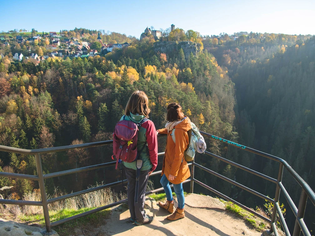 View of Hohnstein Castle from the Hockstein viewpoint Zwei Personen mit Rucksäcken stehen an einem Aussichtspunkt und blicken auf einen bewaldeten Hang mit einem Dorf im Hintergrund bei klarem Wetter.