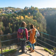 View of Hohnstein Castle from the Hockstein viewpoint Zwei Personen mit Rucksäcken stehen an einem Aussichtspunkt und blicken auf einen bewaldeten Hang mit einem Dorf im Hintergrund bei klarem Wetter.