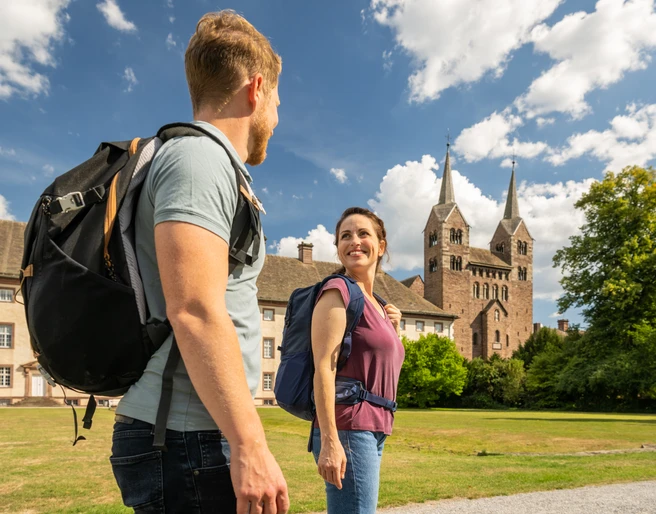 Hoexter-Corvey-Teutoburger-Wald-Tourismus-D-Ketz-074.jpg Ein Paar mit Rucksäcken spaziert lächelnd vor einer historischen Kathedrale mit zwei Türmen.