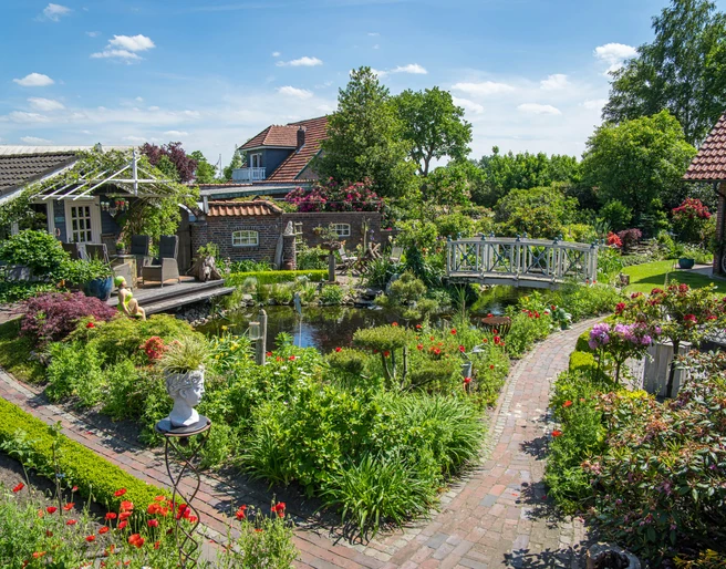 Garten_Privatgarten_Mueller Bollenhagen-32.jpg Ein idyllischer Garten mit blühenden Blumen, einem Teich und einer weißen Brücke.