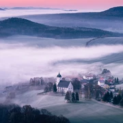 Sonnenaufgang auf dem Papststein mit Blick nach Papstdorf  Nebliges Tal mit einer weißen Kirche und umliegenden Häusern, umgeben von Feldern und Hügeln im Morgenlicht, unter einem rosa Himmel.