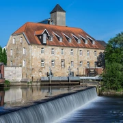 Rheine_Emswehr 2_Foto_ Rheine Tourismus Veranstaltungen eV_Y Mokdad.jpg Historisches Gebäude mit rotem Ziegeldach und Emswehr im Vordergrund bei sonnigem Wetter in Rheine.